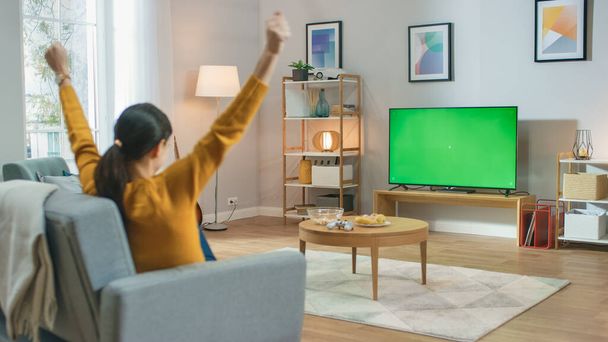 Girl Sitting At Home Sitting on a Couch, Watching Green Chroma Key Screen, Does Winning Gesture with Arms. Man in a Cozy Room Watching Sports Match, News, Sitcom TV Show or a Movie. - Foto, Imagen