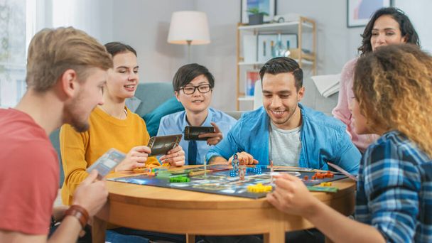 Diverse Group of Guys and Girls Playing in a Strategic Board Game with Cards and Dice. Cozy Living Room in a Daytime - Photo, image