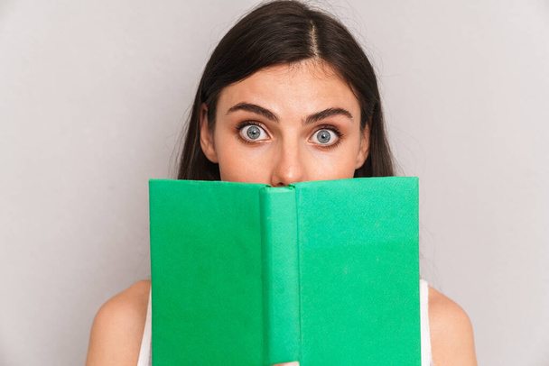 Image closeup of happy brunette woman wearing dress looking at camera and covering her face with book isolated over gray background - Photo, image