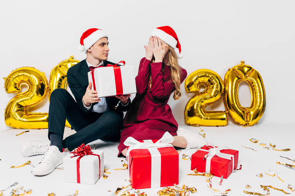 A young Christmas family, a guy and a girl in Santa hats, give gifts sitting on a white background - Foto, Imagen
