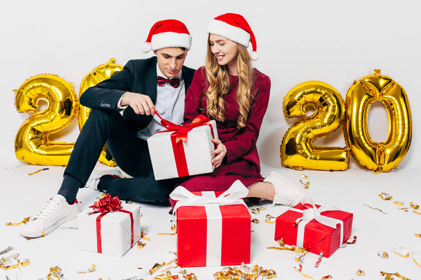 A young Christmas family, a guy and a girl in Santa hats, are opening Christmas presents while sitting on a white background - Foto, Imagen