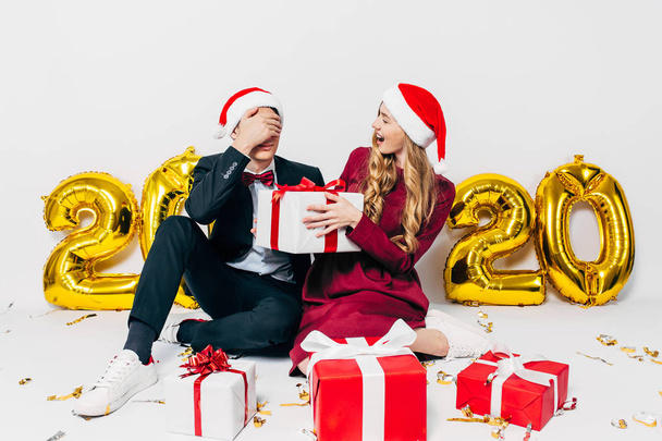 A young Christmas family, a guy and a girl in Santa hats, give gifts sitting on a white background - Foto, Imagen