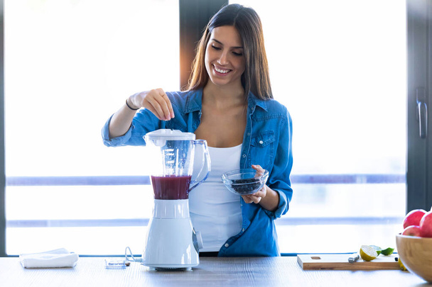 Pretty young woman preparing fruit beverage in blender for fresh breakfast in the kitchen at home. - Photo, image