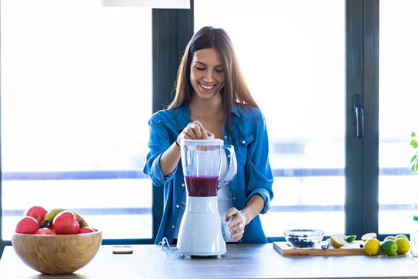Pretty young woman mixing fruit in blender for fresh breakfast in the kitchen at home. - Φωτογραφία, εικόνα