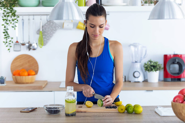 Sporty young woman cutting limes while listening to music in the kitchen at home - Zdjęcie, obraz