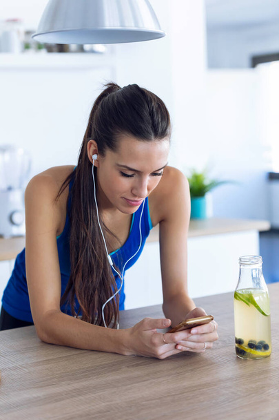 Sporty young woman listening to music with mobile phone after training in the kitchen at home. - Foto, Bild