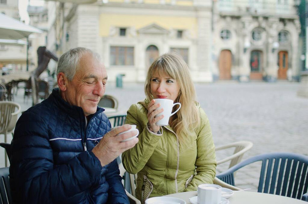 Portrait of happy romantic couple with age difference drinking coffee in cafe with terrace outdoors in the ancient city in the morning during early spring or autumn. - Photo, Image