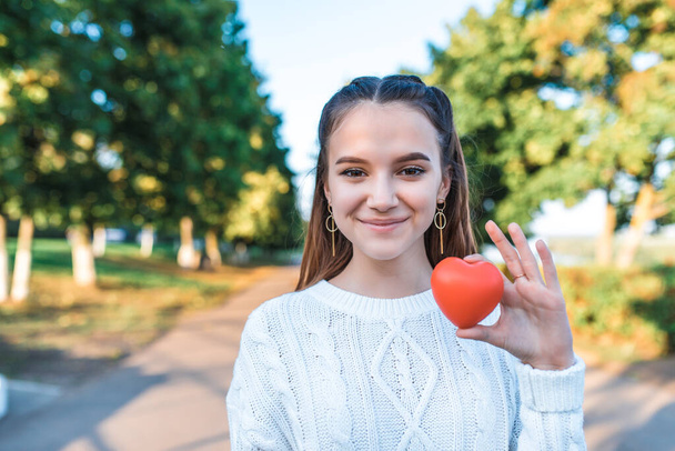 Happy cheerful teenage girl 13-15 years old, summer autumn spring in park, red heart in her hand. Free space text. Concept give love, donation wish of love, help children, save life, positive joy. - Photo, image