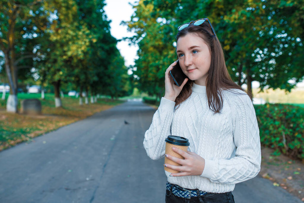teenager girl 12-15 years old, in summer in city park in fall, makes phone call listens to message in online application on Internet. In sweater, cup of coffee tea drink. Free space for text. - Foto, Imagem