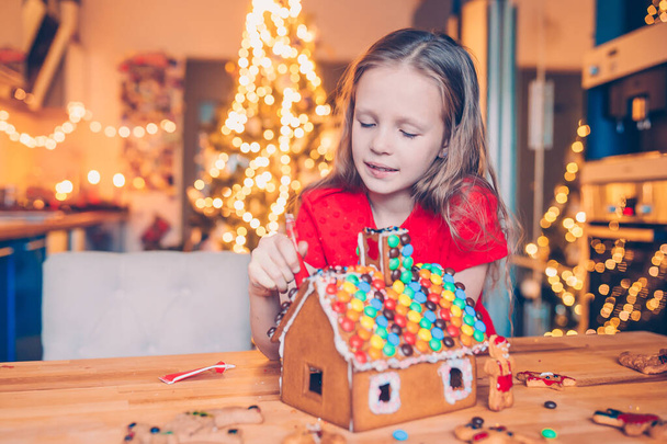 Маленькие девочки делают рождественский пряничный домик у камина в украшенной гостиной. - Фото, изображение