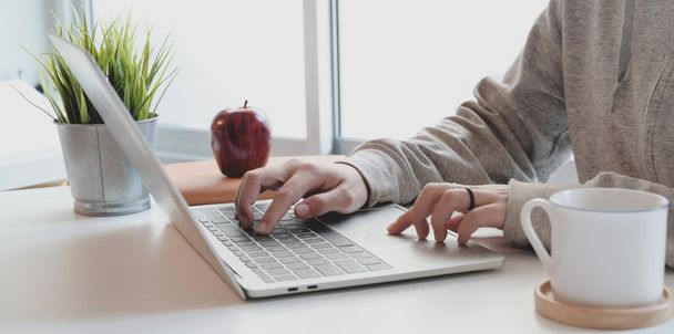 Vista de cerca de la joven mujer escribiendo en el ordenador portátil en un cómodo espacio de trabajo cerca de las ventanas con taza de café
 - Foto, imagen