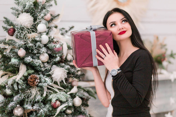 Красива дівчина в чорній сукні і мрійливий вигляд з новорічним подарунком біля ялинки
 - Фото, зображення