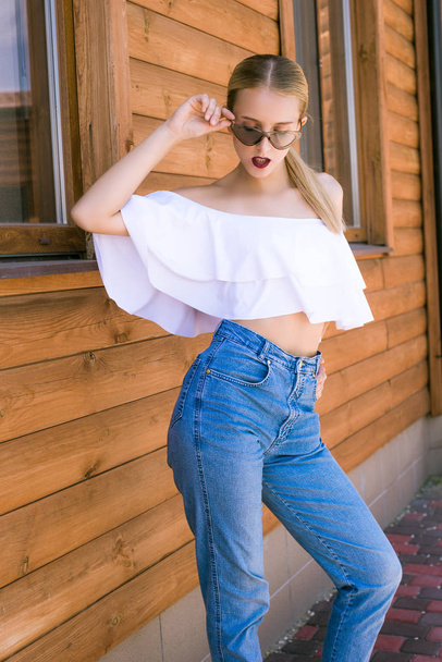 Attractive young girl with a perfect figure and blond hair in trendy jeans and a white top with ruffles and sunglasses in the summer against a wall of wood - Photo, image