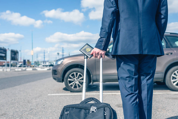 Άντρας με μπλε κοστούμι με βαλίτσα και αμερικάνικο διαβατήριο στο πάρκινγκ του αεροδρομίου με φόντο το αυτοκίνητό του. Σχέδιο επαγγελματικού ταξιδιού - Φωτογραφία, εικόνα