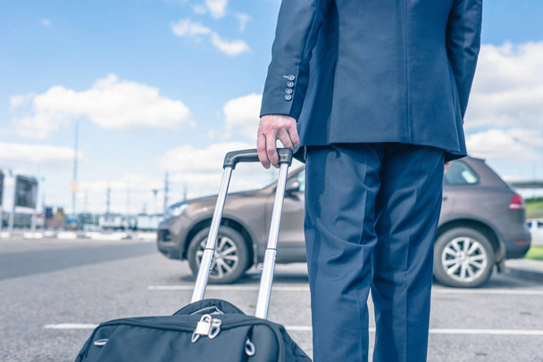 Man in a blue suit with a suitcase at the airport parking on the backdrop of his car. Business trip concept - Фото, зображення