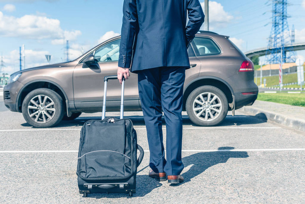 Man in a blue suit with a suitcase at the airport parking on the backdrop of his car. Business trip concept - Fotoğraf, Görsel