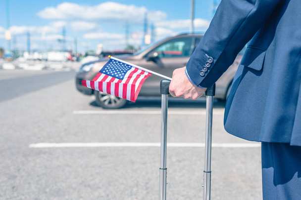 Man in a blue suit with a suitcase and american flag at the airport parking on the backdrop of his car.  concept - Zdjęcie, obraz