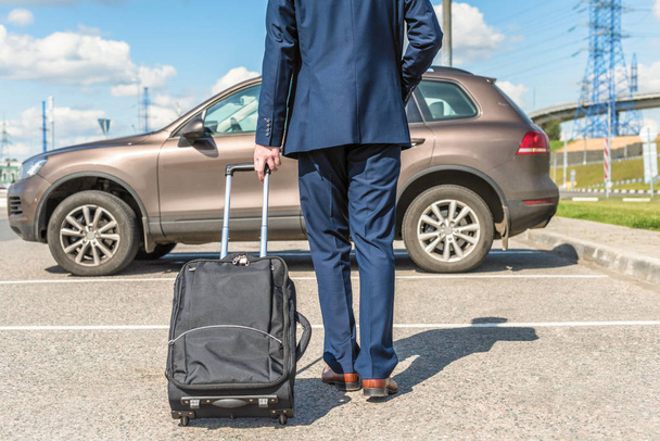 Man in a blue suit with a suitcase at the airport parking on the backdrop of his car. Business trip concept - Fotoğraf, Görsel