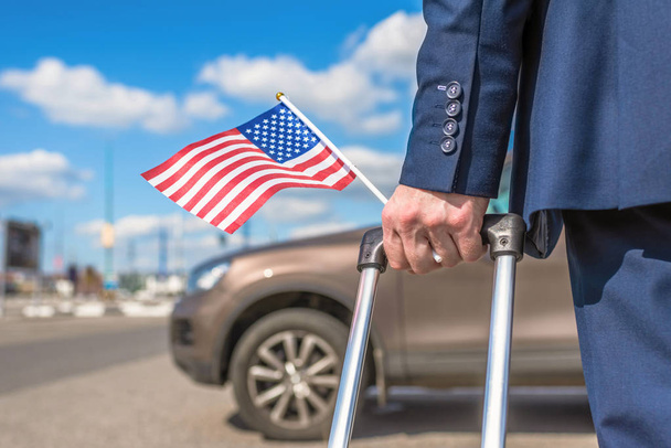 Man in een blauw pak met een koffer en Amerikaanse vlag op het vliegveld parkeren op de achtergrond van zijn auto. concept - Foto, afbeelding