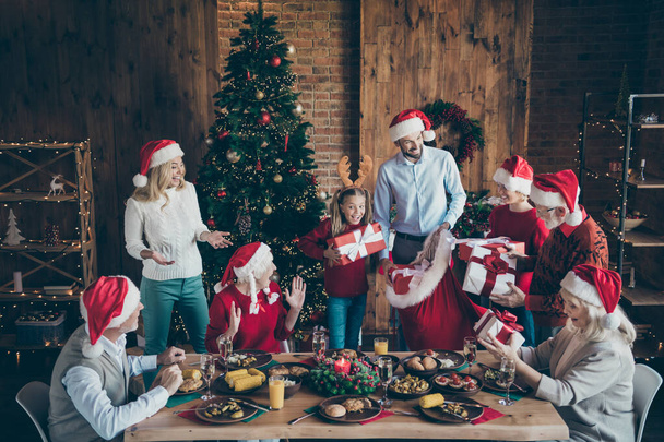 Фото большой семьи с маленькими детьми старший пенсионер сидеть за столом на празднование рождественской трапезы в шляпе Санта-Клауса получить желаемый подарок коробки дома имеющие дух атмосферы X-mas дерево
 - Фото, изображение