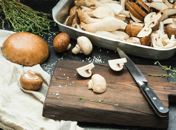 Various raw mushrooms - King trumpet mushroom (Eringi), white and brown mushrooms, Portobello on a dark table with a cutting board and herbs. Selective focus. - Photo, Image