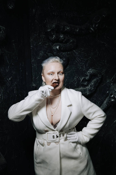 Fiatal torzszülött plusz méretű albínó nő mohón eszik és szereti a csokoládét fekete háttérrel. göndör rövid haj lány fehér ruhában. - Fotó, kép