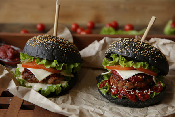 Dva černý sendvič, hamburger nebo burger s hovězím nebo kuřecím masem, rajčata, cibule, sýr, salát a sezamová houska na papíře na dřevěné rustikální desce. Koncept rychlého občerstvení - Fotografie, Obrázek