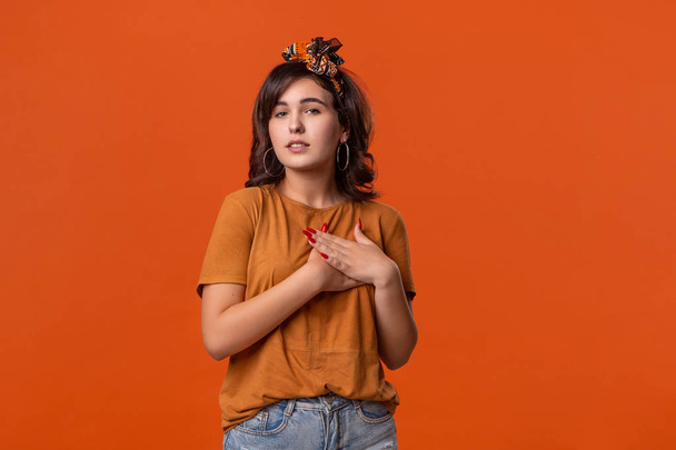Pretty brunette woman in a t-shirt and beautiful headband expressing sincere feeling putting hand to heart standing isolated over orange background. Concept of sincerity - Photo, Image
