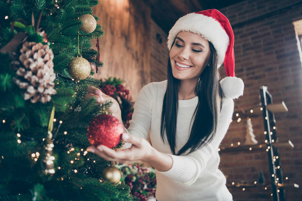 Portrait of positive cheerful girl in santa claus headwear hang christmas ball on evergreen tree for x-mas newyear celebration in house with festive advent decoration ornament indoors - Photo, image