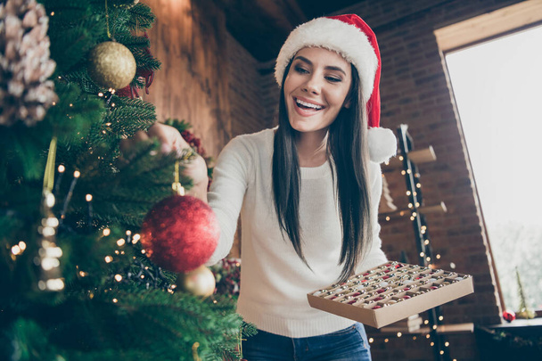 Portrait of positive cheerful girl in santa claus headwear prepare evergreen fir tree for christmas celebration enjoy x-mas tradition wear white sweater in house indoors - Foto, Imagem