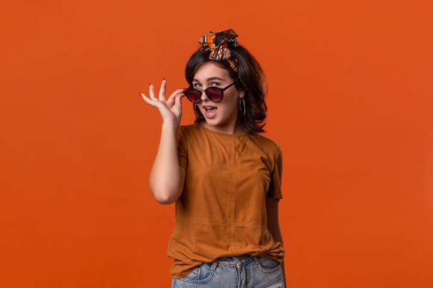 Bella donna bruna in t-shirt e bellissimo cerchietto che esprime emozione di sorpresa tenendo gli occhiali da sole in piedi isolati su sfondo arancione
. - Foto, immagini