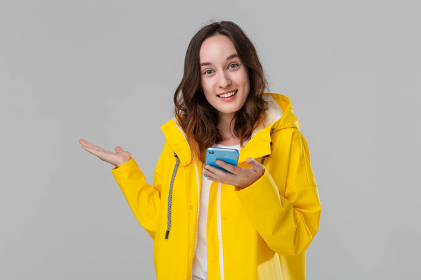 Pretty brunette woman in a yellow raincoat is excited reading notification on the smartphone isolated over grey background. Concept of communication. - Photo, Image