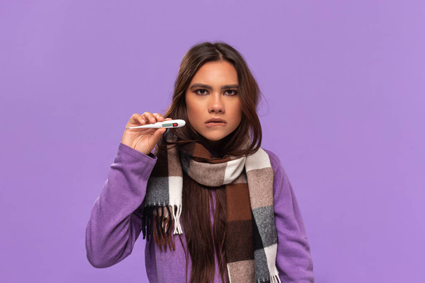 セーターを着たかなりアフリカ系アメリカ人の女の子と居心地の良いチェックスカーフは、紫色の背景に隔離された立って温度計をチェックします。冬に寒さをキャッチ. - 写真・画像