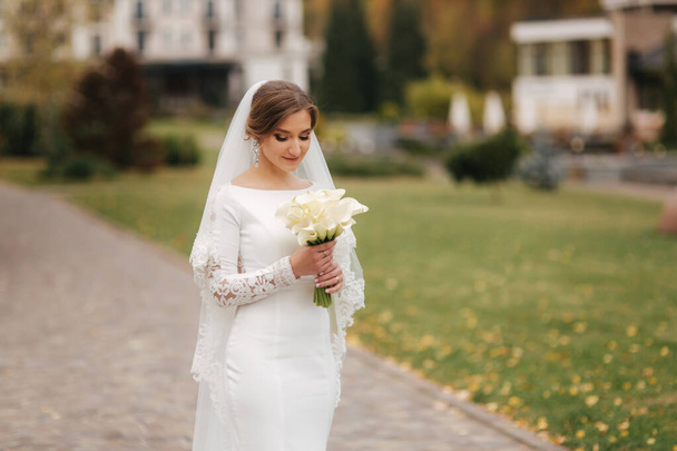 エレガントなウェディングドレスを着た美しい花嫁が公園に立って花の花束を持っています。魅力的な女性の肖像 - 写真・画像