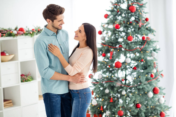Portrait of two people romantic spouses with brunet hair hugging enjoy christnas time x-mas vacation wearing blue denim jeans shirt pastel sweater in house with newyear decoration indoors - Zdjęcie, obraz