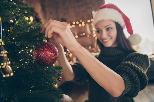 Close up photo of lovely positive girl in santa claus headwear prepare for noel celebration party hand big red balls on christmas evergreen tree in house with newyear decoration indoors - Fotoğraf, Görsel