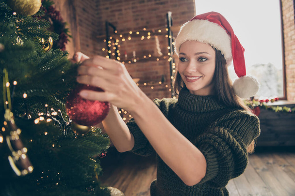 Close up photo of positive cheerful girl in santa claus cap knitted sweater enjoy christmas celebration hang red big bauble on evergreen tree in house with newyear decoration indoors - Foto, Imagem