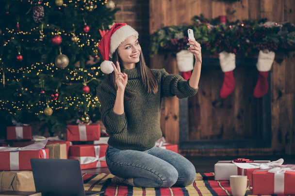 Full size profile side photo of positive woman in santa claus hat have christmas time vacation celebrate take selfie make v-signs sit floor plaid blanket with x-mas decoration lights in house indoors - Foto, Imagem