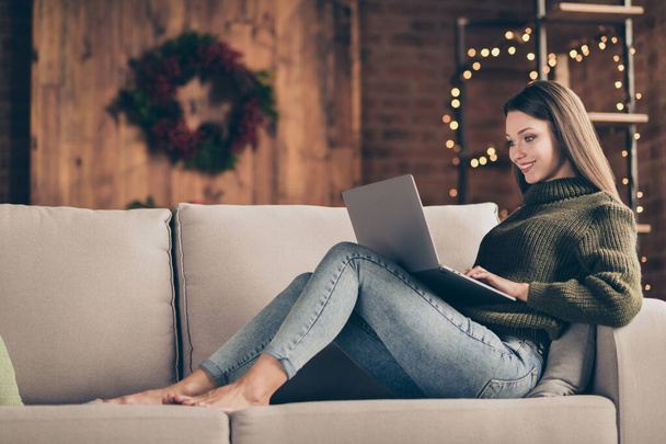 Full length profile side photo of charming positive girl enjoy her christmas time vacations lying on divan type email to her friends send congratulations with newyear use laptop in house indoors - Foto, Bild