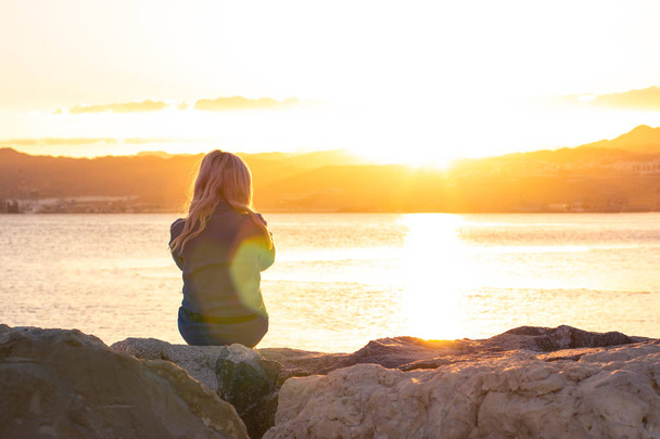samotność uczucie siedzi dziewczyna na kamiennej linii brzegowej w pobliżu morza nabrzeża w jasny pomarańczowy zachód słońca czas z glares humorzasty plakat fotograficzny z pustym miejscu kopii tekstu - Zdjęcie, obraz