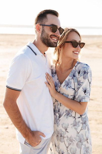Photo of smiling young couple man and woman looking aside and hugging while walking on sunny beach - Foto, Imagem
