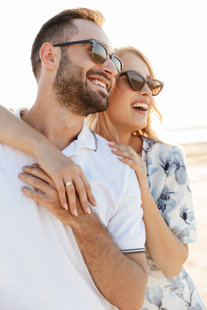 Photo of laughing attractive couple wearing sunglasses looking aside and hugging while walking on sunny beach - Fotografie, Obrázek