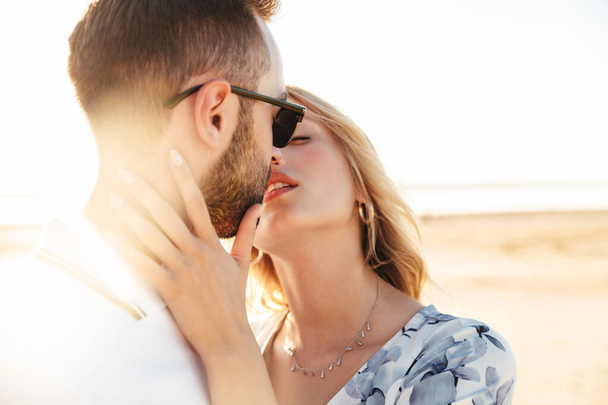 Photo of romantic beautiful couple wearing summer clothes kissing and hugging while walking on sunny beach - Фото, изображение