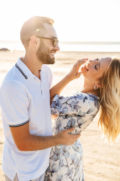 Photo of delighted pretty couple laughing and hugging while walking on sunny beach - Foto, afbeelding