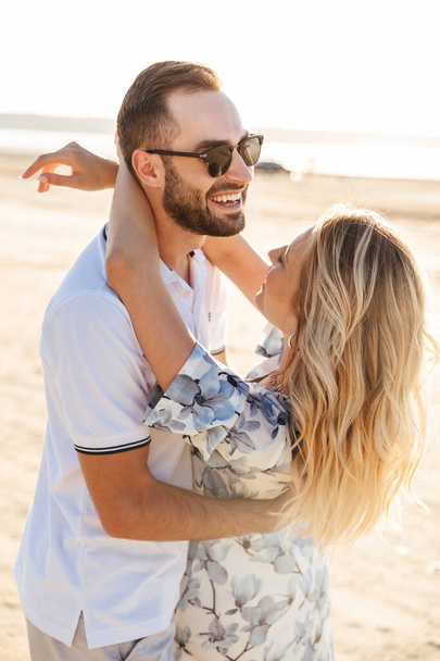 Photo of attractive happy couple laughing and hugging while walking on sunny beach - Foto, Imagen