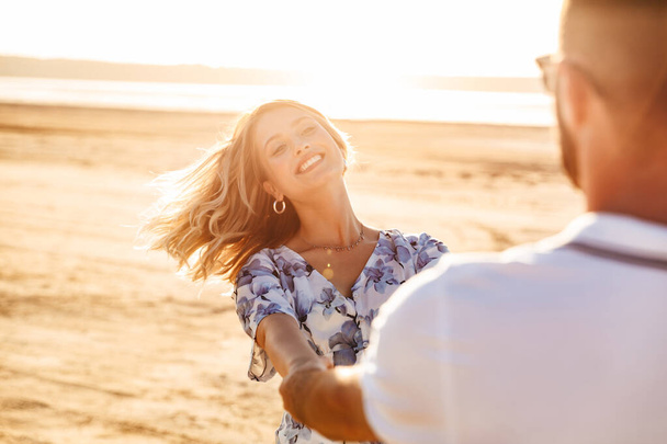 Zdjęcie pięknej radosnej pary uśmiechającej się i wirującej podczas spaceru po słonecznej plaży - Zdjęcie, obraz