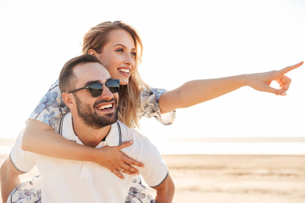Photo of unshaven cheerful man smiling and giving piggyback ride seductive woman while walking on sunny beach - Photo, image