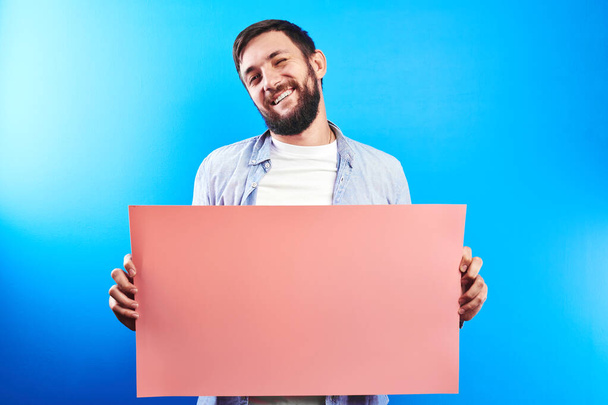 Handsome bearded caucasian man holding blank pink poster board in hands with copy space for text or advertising inscription isolated in blue studio - Fotografie, Obrázek