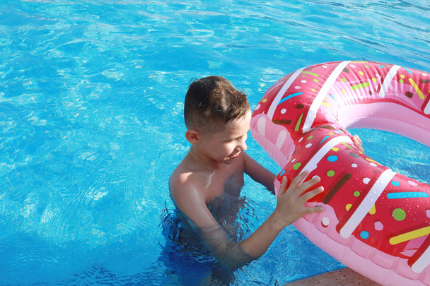 Ευτυχισμένο χαριτωμένο αγόρι στην πισίνα σε μια ηλιόλουστη μέρα σε ένα στρώμα αέρα με τη μορφή ενός ροζ ντόνατ - Φωτογραφία, εικόνα