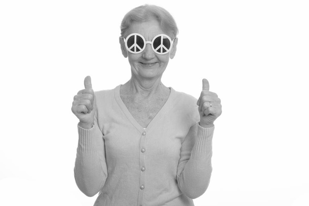 glückliche Seniorin lächelt, während sie Sonnenbrille mit Friedenszeichen trägt und Daumen hoch gibt - Foto, Bild